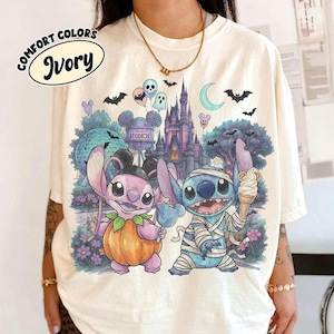 May include: An ivory t-shirt featuring a graphic of two blue and purple alien characters in Halloween costumes. One character wears a pumpkin suit, the other is wrapped in bandages holding an ice cream cone. The background shows a castle, a geodesic dome, and bats.