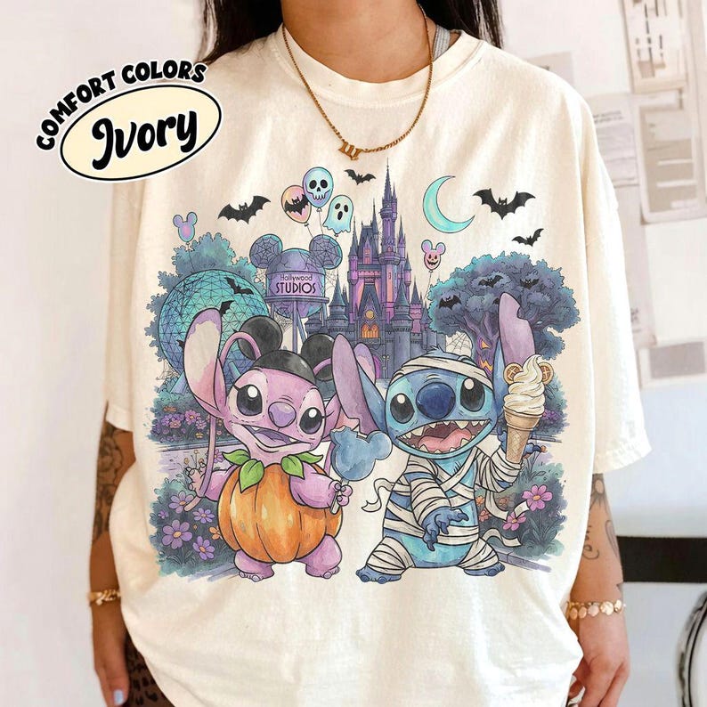 May include: An ivory t-shirt featuring a graphic of two blue and purple alien characters in Halloween costumes. One character wears a pumpkin suit, the other is wrapped in bandages holding an ice cream cone. The background shows a castle, a geodesic dome, and bats.
