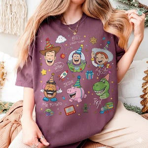 May include: A maroon t-shirt featuring cartoon illustrations of characters from the movie Toy Story, including Woody, Buzz Lightyear, Jessie, Mr. Potato Head, Rex, and Hamm. The shirt has the text "Buzz Lightyear" and character names.