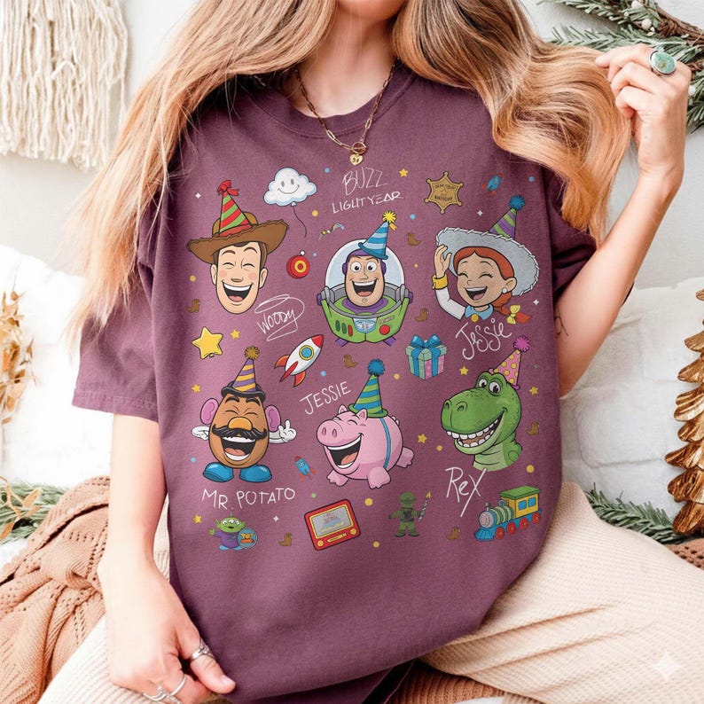 May include: A maroon t-shirt featuring cartoon illustrations of characters from the movie Toy Story, including Woody, Buzz Lightyear, Jessie, Mr. Potato Head, Rex, and Hamm. The shirt has the text "Buzz Lightyear" and character names.