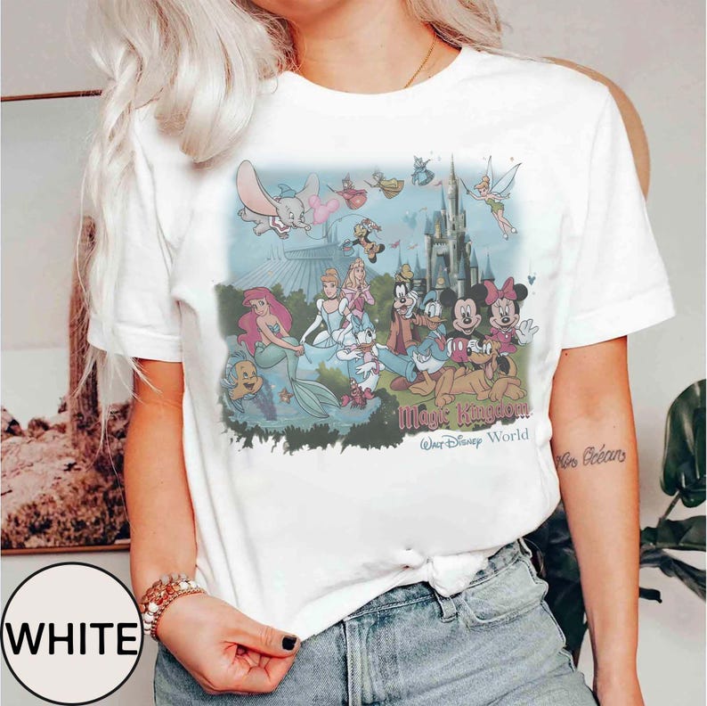 K&ouml;nnte beinhalten: Wei&szlig;es T-Shirt mit einer farbenfrohen Grafik von Disney-Figuren, darunter Arielle, Mickey Mouse und Cinderella, vor einem Schloss. Der Text "Magic Kingdom Walt Disney World" ist unter der Grafik aufgedruckt.