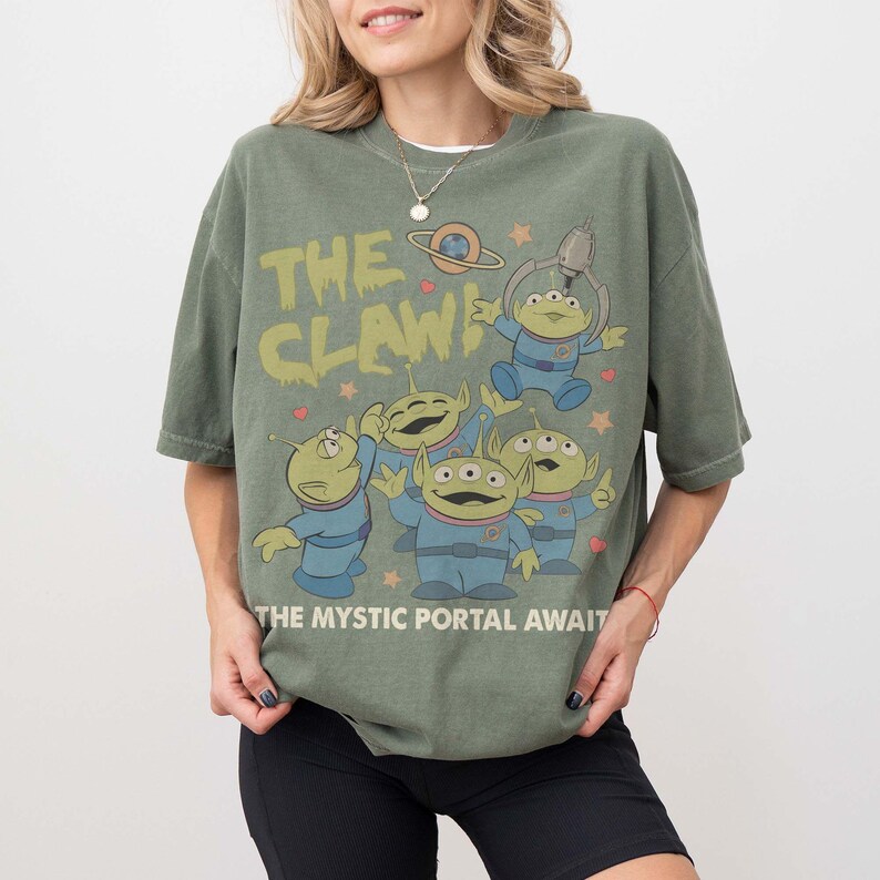 May include: Olive green t-shirt featuring the phrase "THE CLAW!" above a graphic of the Little Green Men from Toy Story. The text "THE MYSTIC PORTAL AWAITS" is printed below the graphic. The shirt has short sleeves.