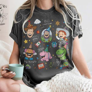 May include: Dark gray t-shirt featuring cartoon characters Woody, Buzz Lightyear, Jessie, Mr. Potato Head, and Rex. The shirt has a distressed look and includes the text "Buzz Lightyear" and character names.