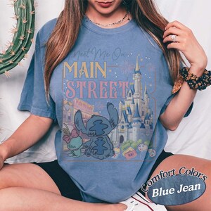 May include: Blue jean t-shirt with a graphic featuring Stitch and a castle, with the text "Meet Me On Main Street." The design includes fireworks and a ticket. The words "Comfort Colors" are also visible.