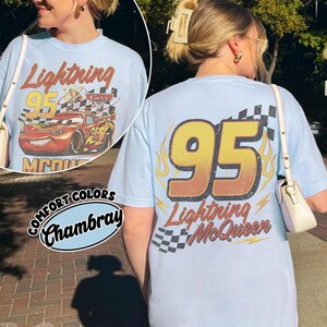 Może przedstawiać: Jasnoniebieska koszulka z Lightning McQueen z filmu Auta. Prz&oacute;d przedstawia numer 95 i logo Cars, a tył duży 95 z płomieniami i "Lightning McQueen". Koszulka jest oznaczona jako "Comfort Colors Chambray".