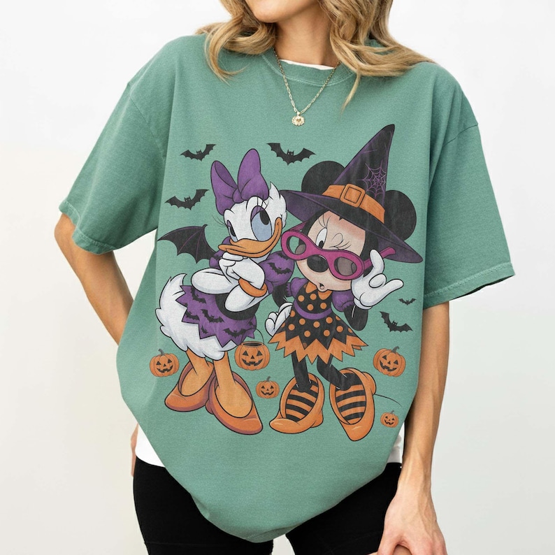 Może przedstawiać: Szałwiowo-zielony t-shirt z kr&oacute;tkim rękawem z nadrukiem graficznym przedstawiającym dwie postacie z kresk&oacute;wek w kostiumach na Halloween. Jedna postać ma na sobie fioletową sukienkę w nietoperze, a druga kapelusz czarownicy i okulary, w otoczeniu pomarańczowych dyń.