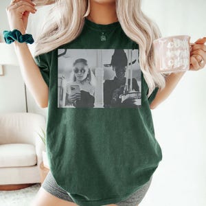 May include: Dark green t-shirt featuring a black and white image of two people. One person is wearing sunglasses and holding a phone, and the other is wearing a witch hat. The shirt is a solid color and has a relaxed fit.
