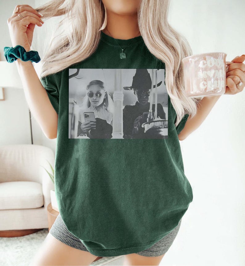 May include: Dark green t-shirt featuring a black and white image of two people. One person is wearing sunglasses and holding a phone, and the other is wearing a witch hat. The shirt is a solid color and has a relaxed fit.
