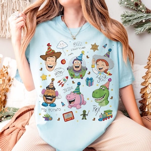 May include: Light blue t-shirt featuring colorful cartoon illustrations of characters from the Toy Story movie. The design includes Woody, Buzz Lightyear, Jessie, Mr. Potato Head, Rex, and Hamm, along with the text "Buzz Lightyear".