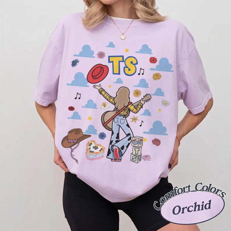May include: A light purple t-shirt featuring a graphic of a person with long hair playing an acoustic guitar, wearing a yellow plaid shirt and cow-print pants. The design includes blue clouds, yellow stars, flowers, a red hat, a brown hat, and blocks with the letters TS.