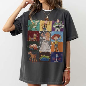 May include: A charcoal gray short-sleeve t-shirt featuring a grid of Toy Story characters including Rex, Woody, Buzz Lightyear, Slinky Dog, Jessie, Mr. and Mrs. Potato Head, Bullseye, the Aliens, Forky, and a central figure of a blonde singer.