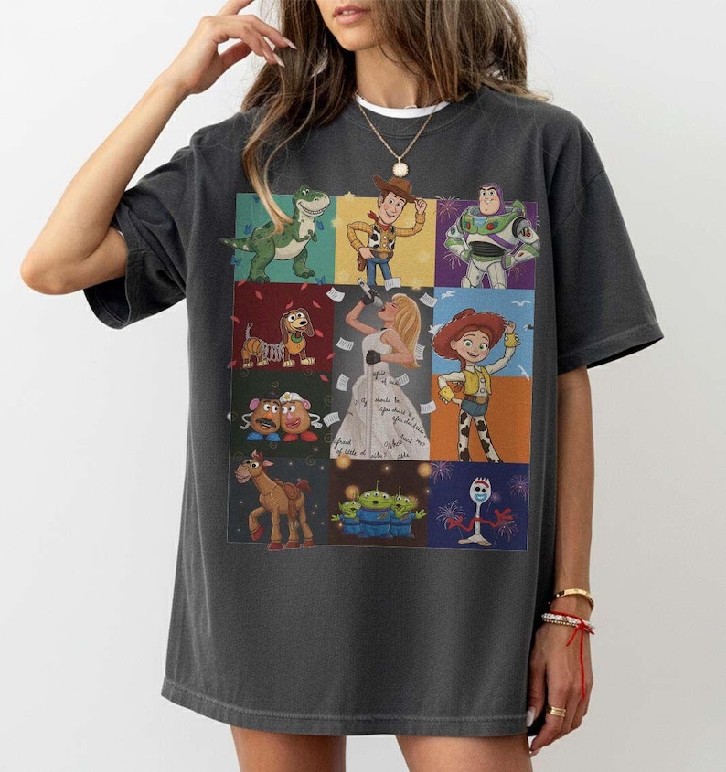 May include: A charcoal gray short-sleeve t-shirt featuring a grid of Toy Story characters including Rex, Woody, Buzz Lightyear, Slinky Dog, Jessie, Mr. and Mrs. Potato Head, Bullseye, the Aliens, Forky, and a central figure of a blonde singer.
