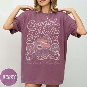 May include: A berry-colored short-sleeve t-shirt featuring a graphic print in white and light pink. The design includes rope-style lettering that reads Cowgirl Like Me, a cowboy hat, a horseshoe, musical notes, and the text Takes One to Know One.