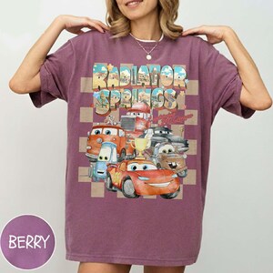 Peut inclure: T-shirt de couleur baie avec un motif des personnages du film Cars et le texte "Radiator Springs" dans un design r&eacute;tro color&eacute;. Le motif comprend divers personnages de voitures et un fond &agrave; carreaux.