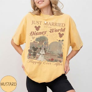 Pu&ograve; includere: T-shirt giallo senape con la scritta "Just Married Disney World Est 2026" e una grafica di Topolino e Minnie in abito da sposi. La maglietta presenta un design in stile vintage con il tema di un matrimonio a Disney World.