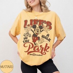 May include: Mustard yellow t-shirt featuring a vintage Mickey Mouse graphic with the text "LIFE'S A WALK IN THE PARK" and "Walt Disney World". The design includes Mickey Mouse with arms outstretched and a castle illustration.