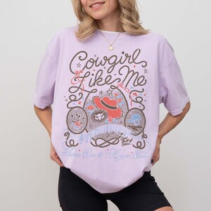 May include: A light purple short-sleeve t-shirt featuring a graphic print with the text Cowgirl like Me, Takes One to Know One, and Eyes full of Stars. The design includes a red cowboy hat, a lasso, a horseshoe, and musical notes in brown and blue.