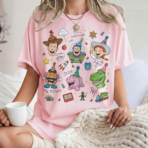 May include: Pink t-shirt featuring colorful cartoon illustrations of characters from the Toy Story movie franchise. The design includes Woody, Buzz Lightyear, Jessie, Mr. Potato Head, Rex, and Hamm, with party hats and other festive elements.