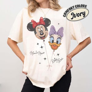 Pu&ograve; includere: Una t-shirt a maniche corte color avorio con una grafica di Minnie Mouse e Daisy Duck raffigurate come palloncini. Il design include il testo Minnie Mouse e Daisy Duck, insieme a piccoli accenti a forma di stella. Etichetta Comfort Colors Ivory.