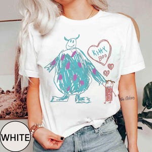May include: White t-shirt featuring a whimsical, hand-drawn design. The graphic includes a large blue monster with purple spots, holding hands with a small red figure. A heart with "Kitty Boo" is above them. The word "WHITE" is in a circle.