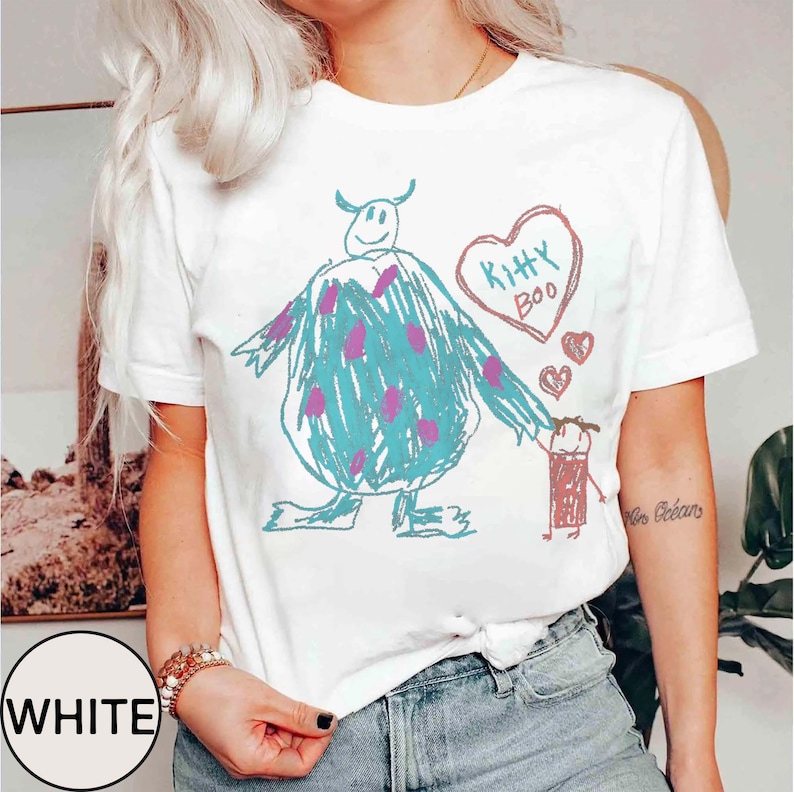 May include: White t-shirt featuring a whimsical, hand-drawn design. The graphic includes a large blue monster with purple spots, holding hands with a small red figure. A heart with "Kitty Boo" is above them. The word "WHITE" is in a circle.
