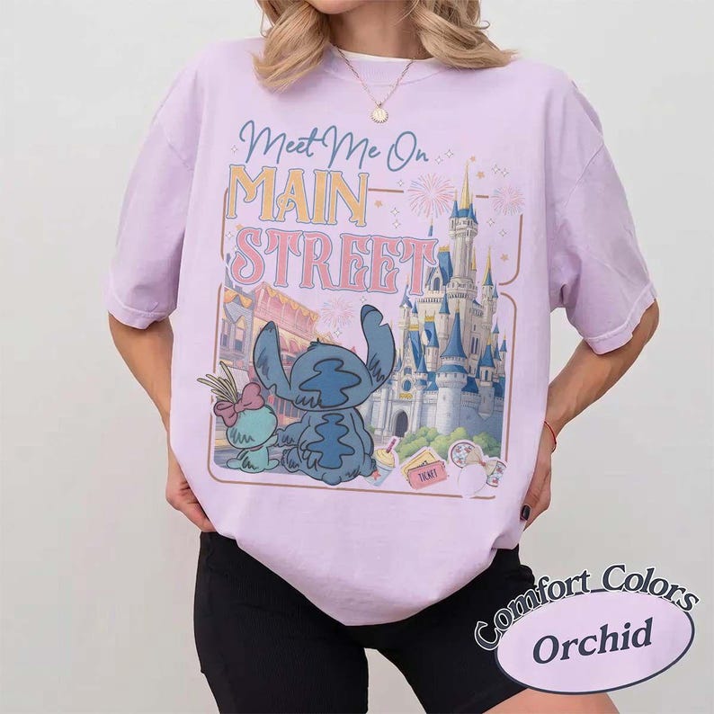May include: A light purple t-shirt with the words "Meet Me On Main Street" and a graphic of Stitch and Angel in front of a castle. The shirt is a Comfort Colors Orchid.