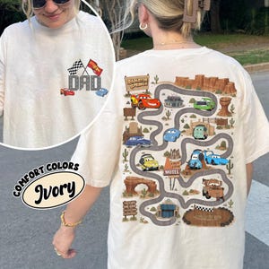 Peut inclure: Un t-shirt ivoire avec un motif frontal composé de deux drapeaux à damier et du texte DAD, et un grand motif au dos représentant une carte routière sinueuse dans un paysage désertique avec des voitures, des cactus et le motel Cozy Cone.