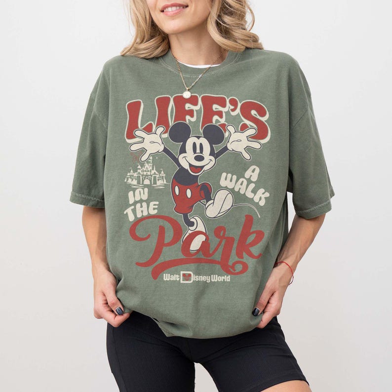 May include: Olive green t-shirt featuring Mickey Mouse with the text "Life's a Walk in the Park" in red and white. The shirt also includes the Walt Disney World logo. The design is vintage-inspired.