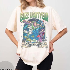 Peut inclure: T-shirt ivoire avec un graphisme de Buzz l'&Eacute;clair de Toy Story. Le motif comprend le texte "Buzz Lightyear" et "Disney Toy Story Laser Blast". Le graphisme est dans les tons de vert, de violet et de bleu.