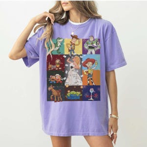 May include: A light purple short-sleeve t-shirt featuring a grid of nine colorful illustrations of various animated characters, including a cowboy, a space ranger, a cowgirl, a dinosaur, a dog, a horse, potato figures, aliens, and a white spork character.