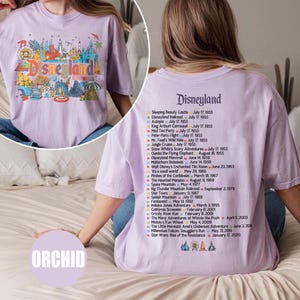 Puede incluir: Camiseta lila con un gr&aacute;fico de Disneyland en la parte delantera y una lista de atracciones en la parte trasera. El gr&aacute;fico frontal incluye la palabra "Disneyland" e iconos del parque. La parte trasera enumera las fechas de apertura de las atracciones.