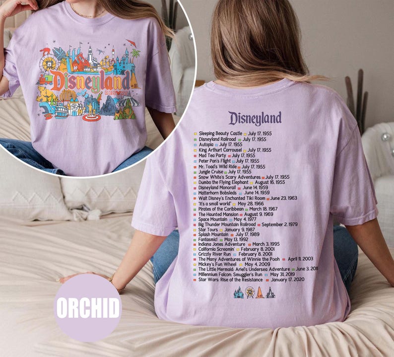 Puede incluir: Camiseta lila con un gr&aacute;fico de Disneyland en la parte delantera y una lista de atracciones en la parte trasera. El gr&aacute;fico frontal incluye la palabra "Disneyland" e iconos del parque. La parte trasera enumera las fechas de apertura de las atracciones.