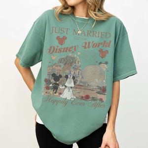 Pu&ograve; includere: T-shirt verde con grafica di Topolino e Minni in abito da sposi, la scritta "Just Married Disney World" e "Happily Ever After". Il design include fuochi d'artificio, palloncini e monumenti di Disney World.