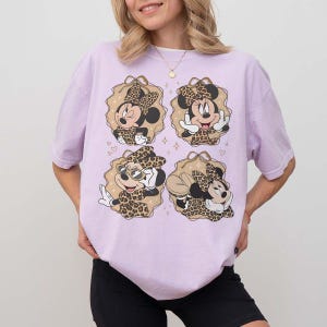 May include: A light purple t-shirt featuring four Minnie Mouse designs with leopard print bows and outfits. Each design is set within a tan-colored, scalloped frame with hearts and stars. The shirt is a casual, oversized style.