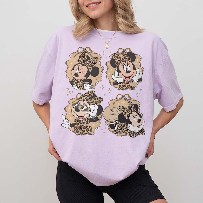 May include: A light purple t-shirt featuring four Minnie Mouse designs with leopard print bows and outfits. Each design is set within a tan-colored, scalloped frame with hearts and stars. The shirt is a casual, oversized style.