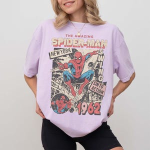 K&ouml;nnte beinhalten: Helllila T-Shirt mit einem Vintage-Spider-Man-Grafik. Das Design enth&auml;lt die W&ouml;rter "THE AMAZING SPIDER-MAN NEW YORK" und "1962". Die Grafik zeigt Spider-Man in Aktion, im Retro-Comic-Stil.