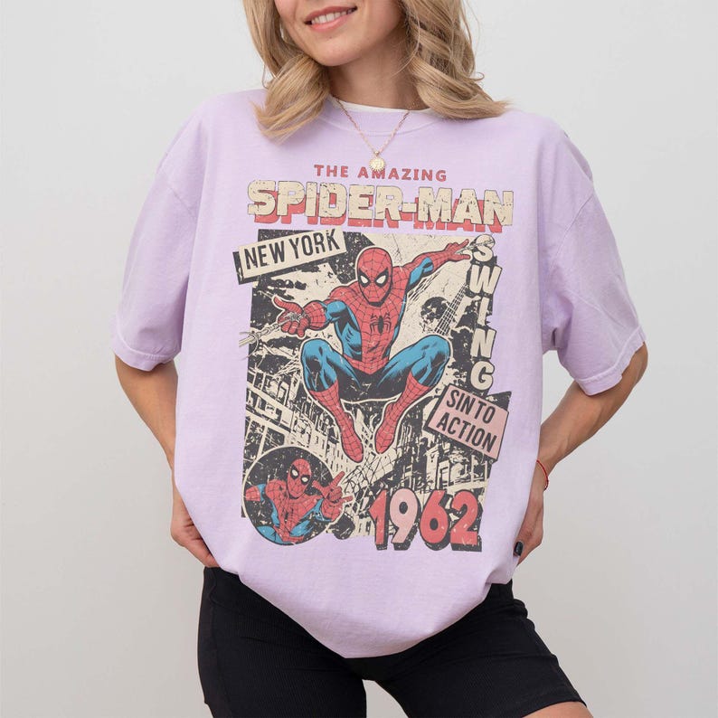 K&ouml;nnte beinhalten: Helllila T-Shirt mit einem Vintage-Spider-Man-Grafik. Das Design enth&auml;lt die W&ouml;rter "THE AMAZING SPIDER-MAN NEW YORK" und "1962". Die Grafik zeigt Spider-Man in Aktion, im Retro-Comic-Stil.