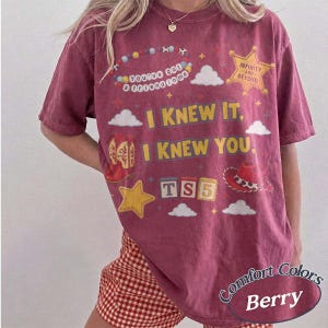 May include: A berry-colored short-sleeve t-shirt featuring a graphic print with the text "I KNEW IT, I KNEW YOU," "You've got a friend in me," "INFINITY AND BEYOND," and "TS5." The design includes illustrations of a cowboy boot, hat, and stars.