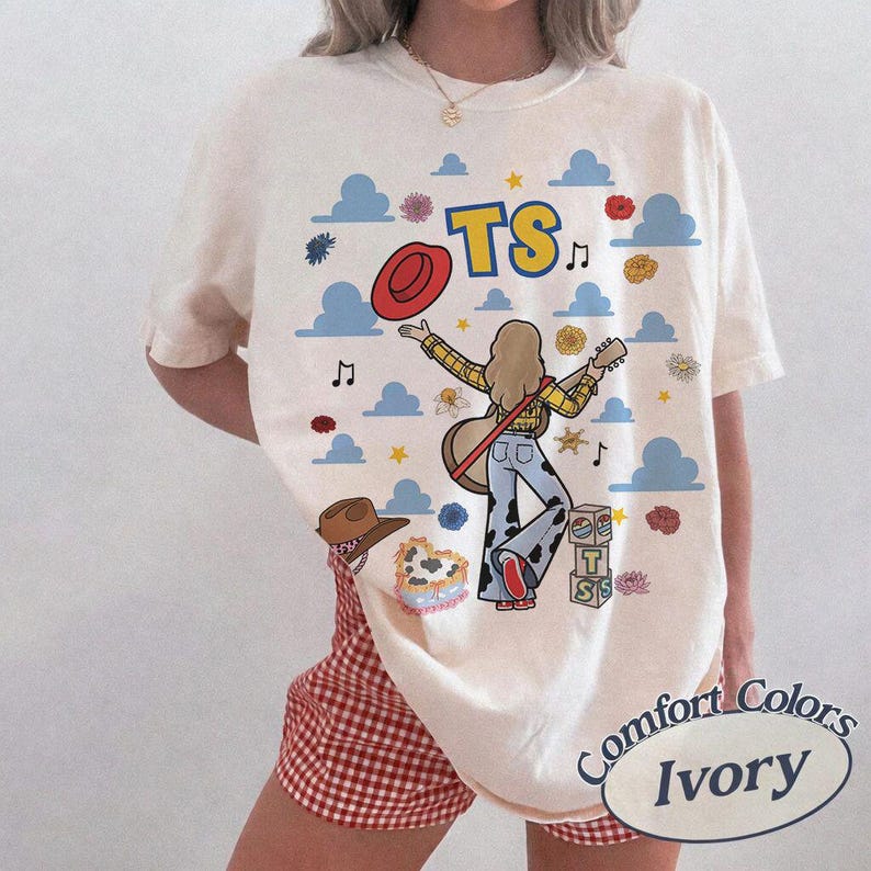 May include: An ivory t-shirt featuring a graphic print of a person with long hair wearing a yellow plaid shirt and cow-print pants, holding a guitar. The design includes blue clouds, red flowers, a red hat, musical notes, and the letters TS.