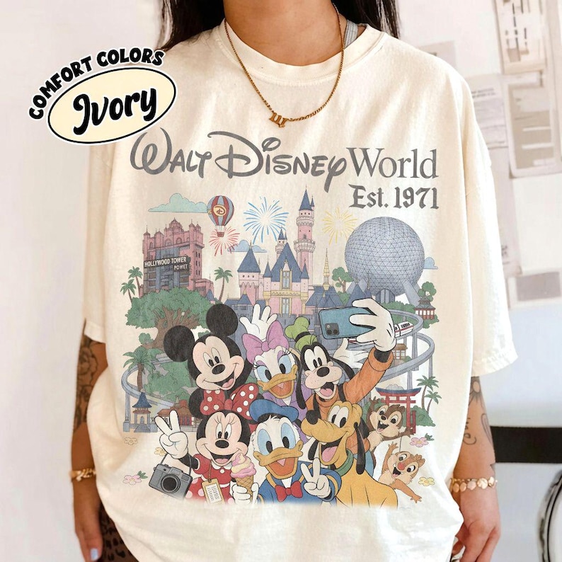 May include: An ivory t-shirt featuring a graphic print of Walt Disney World Est. 1971 with various Disney characters including Mickey Mouse, Minnie Mouse, Donald Duck, Daisy Duck, Goofy, Pluto, Chip, and Dale in front of park landmarks.