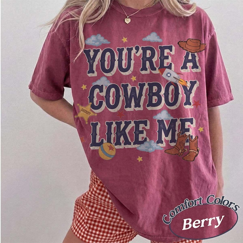 May include: A berry-colored short-sleeve t-shirt featuring the text YOU'RE A COWBOY LIKE ME in dark blue letters. The design includes graphics of a brown cowboy hat, a rocket, a sheriff star, a colorful ball, and brown cowboy boots.