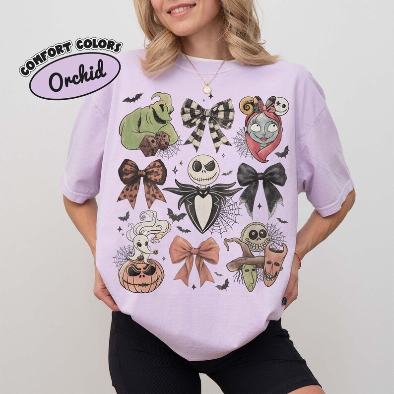 以下が含まれることがあります： オーキッドカラーのTシャツで、『ナイトメアー・ビフォア・クリスマス』のキャラクターとリボンのグラフィックプリントが施されています。ジャック・スケリントン、サリー、ウギー・ブギー、ゼロ、ロック、ショック、バレルが描かれ、黒と白のリボンが散りばめられています。