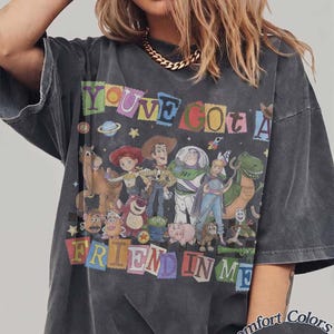 May include: A charcoal gray t-shirt featuring a graphic print of various animated characters from the Toy Story franchise. The text "YOU'VE GOT A FRIEND IN ME" is displayed in colorful, blocky letters above and below the characters. The shirt has a faded, vintage-style wash and short sleeves.
