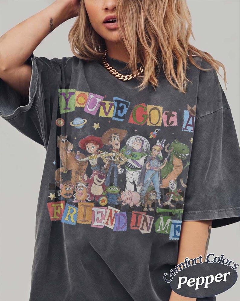 May include: A charcoal gray t-shirt featuring a graphic print of various animated characters from the Toy Story franchise. The text "YOU'VE GOT A FRIEND IN ME" is displayed in colorful, blocky letters above and below the characters. The shirt has a faded, vintage-style wash and short sleeves.