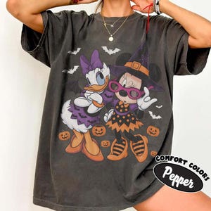 Może przedstawiać: Ciemnoszary t-shirt z kr&oacute;tkim rękawem z grafiką przedstawiającą Daisy Duck i Minnie Mouse w kostiumach na Halloween. Daisy ma na sobie fioletową sukienkę w nietoperze, a Minnie nosi kapelusz czarownicy, okulary i pomarańczową sukienkę w kropki.