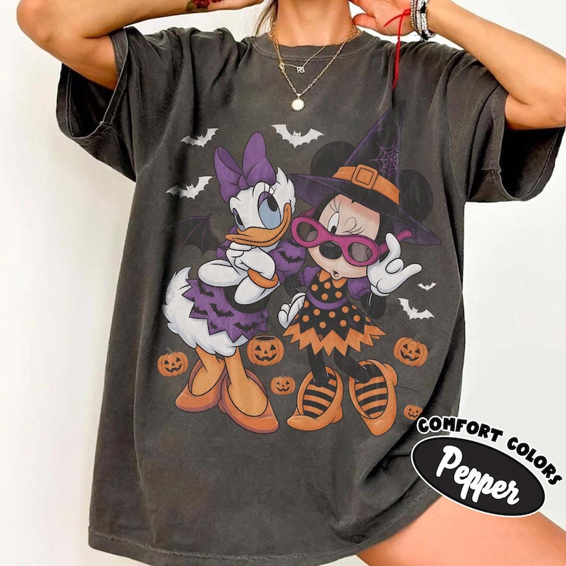 Może przedstawiać: Ciemnoszary t-shirt z kr&oacute;tkim rękawem z grafiką przedstawiającą Daisy Duck i Minnie Mouse w kostiumach na Halloween. Daisy ma na sobie fioletową sukienkę w nietoperze, a Minnie nosi kapelusz czarownicy, okulary i pomarańczową sukienkę w kropki.