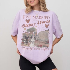Pu&ograve; includere: Maglietta lilla con grafica Disney World con Topolino e Minnie. La maglietta ha la scritta "Just Married Disney World Happily Ever After". Il design include un castello e Spaceship Earth.
