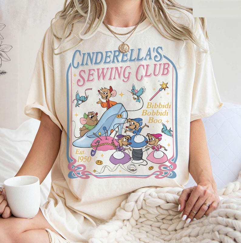 May include: Cream-colored t-shirt featuring a whimsical design of Cinderella's Sewing Club. The graphic includes a blue glass slipper, mice, sewing tools, and the text "Cinderella's Sewing Club Est. 1950" in a retro style.