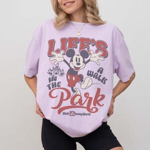May include: A light purple t-shirt featuring a graphic of Mickey Mouse with the text "Life's a Walk in the Park" and "Walt Disney World". The design includes a castle illustration and retro font.