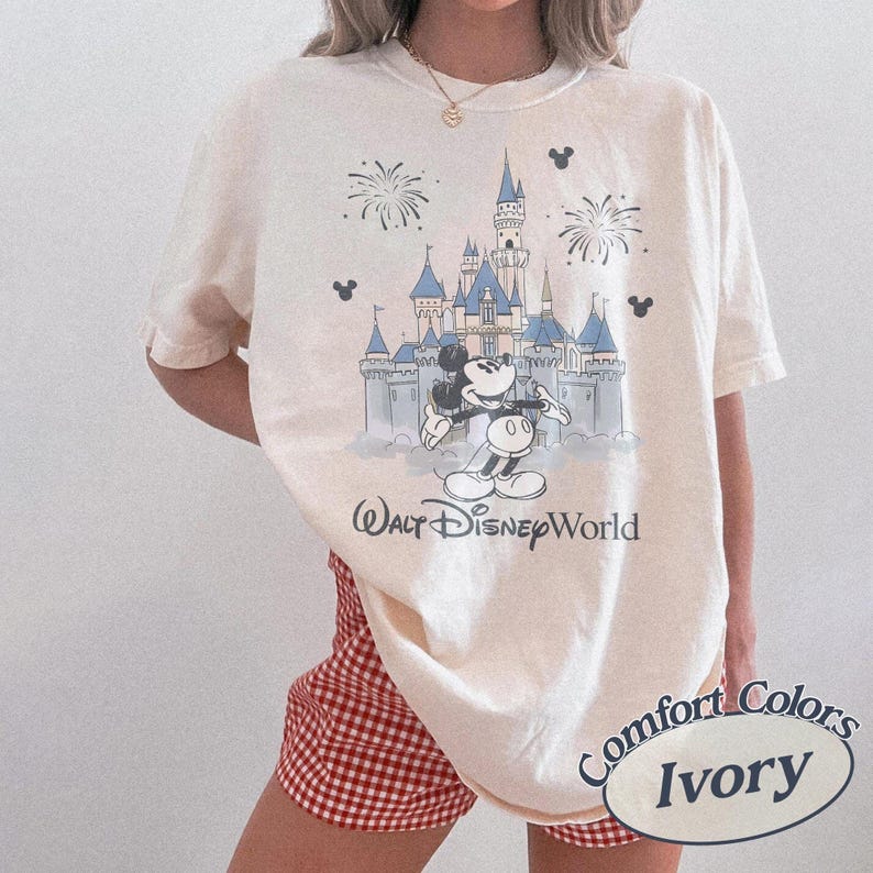 May include: Ivory t-shirt with a Walt Disney World graphic featuring Mickey Mouse and Cinderella's castle. The design includes fireworks and Mickey Mouse silhouettes. The shirt is labelled "Comfort Colors Ivory".